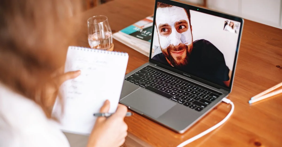 Un homme avec un masque facial en appel vidéo avec une femme qui prend des notes devant un ordinateur portable.